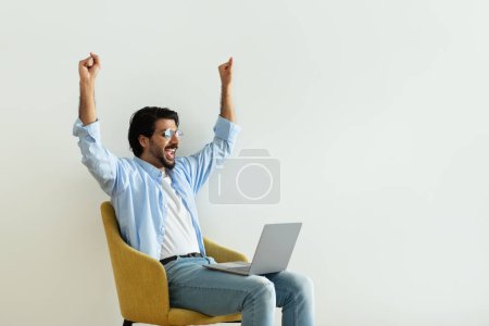 Téléchargez les photos : Heureux heureux millénariste Moyen-Orient gars dans des lunettes lève les mains, célébrer la victoire, faire un geste de succès avec ordinateur portable sur chaise dans la chambre sur fond de mur blanc, profiter de gagner, annonce et offre - en image libre de droit