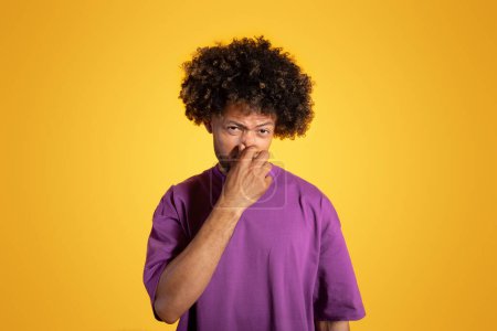 Foto de Desesperado triste maduro negro rizado hombre en púrpura camiseta cubre la nariz con la mano, sufre de mal olor aislado en el fondo amarillo, estudio. Apestoso, problemas de higiene, anuncio y oferta, emociones humanas - Imagen libre de derechos
