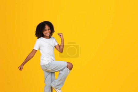 Téléchargez les photos : Jeune fille bouclée souriante en t-shirt blanc dansant, s'amuse seule, isolée sur fond de studio jaune. Temps libre pour les élèves, mode de vie, études, connaissances et éducation à l'école - en image libre de droit