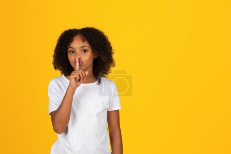 Téléchargez les photos : Joyeux mystérieux adolescent fille bouclée en t-shirt blanc faire un geste shh, signe secret, fond isolé studio jaune. commérages, bonnes nouvelles, étude scolaire, émotions des élèves - en image libre de droit
