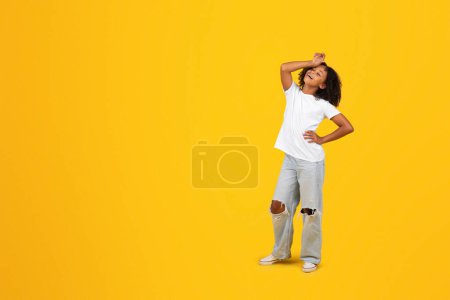 Photo for Cheerful tired adolescent black curly girl in white t-shirt wipes sweat from forehead, isolated yellow studio background. Difficulties in education, knowledge, study, emotions, prepare to exam - Royalty Free Image