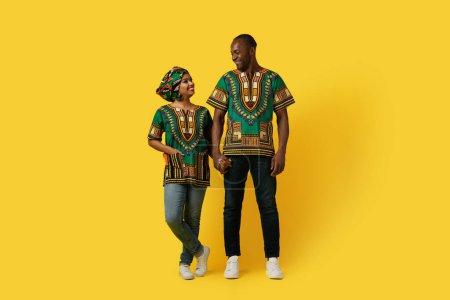 Photo for Loving happy bright black couple wearing national african shirts holding hands, looking at each other and smiling on yellow studio background, full length, copy space. Relationships, family concept - Royalty Free Image