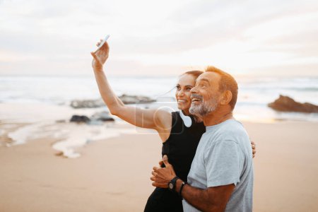 Photo for Positive mature european family in sportswear rest from training, taking selfie on smartphone in morning on sea beach, outdoor. Social networks, video call, sports, fitness and body care - Royalty Free Image