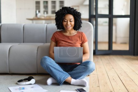 Téléchargez les photos : Concept freelance. Heureuse femme noire utilisant un ordinateur portable pour le travail à distance à la maison, Souriant jeune afro-américaine pigiste femelle assise sur le sol dans le salon et tapant sur l'ordinateur, Copier l'espace - en image libre de droit