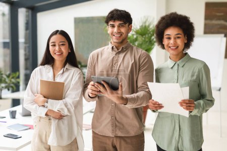 Photo for Business Diversity. Asian, Middle Eastern And African American Coworkers Holding Digital Tablet And Papers Smiling To Camera Standing In Office. Shot Of Successful Young Coworkers Trio. Teamwork - Royalty Free Image