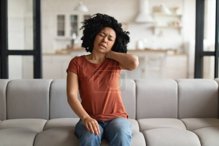 Foto de Mujer negra cansada que sufre dolor de cuello mientras está sentada en el sofá en casa, mujer afroamericana agotada que masajea el área dolorosa y el ceño fruncido, que sufre osteoporosis o artritis, espacio copiado - Imagen libre de derechos