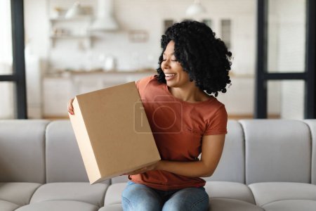 Téléchargez les photos : Concept de livraison. Heureuse femme noire tenant une grande boîte en carton à la maison, jeune femme afro-américaine joyeuse regardant le colis livré et souriant, assise sur le canapé dans le salon - en image libre de droit