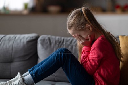 Photo for Upset little girl sitting on couch at home and crying, depressed female child covering face with hands and sobbing, feeling lonely or insulted after quarrel with parents, side view with copy space - Royalty Free Image