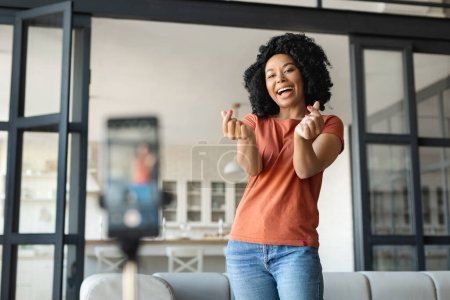 Photo for Cheerful Black Woman Making Finger Heart Gesture At Camera While Recording Video At Home, Happy Young African American Lady Capturing Content For Social Networks, Using Smartphone On Tripod - Royalty Free Image