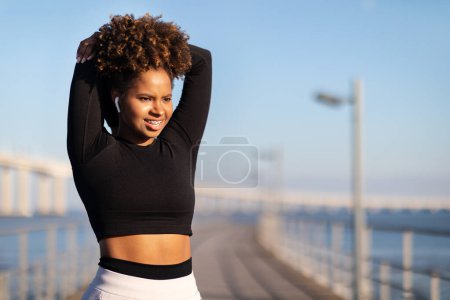 Foto de Retrato de atlética mujer negra en ropa deportiva haciendo ejercicio al aire libre, sonriendo joven mujer afroamericana calentándose antes de correr, estirando los músculos del brazo y mirando el espacio de copia - Imagen libre de derechos