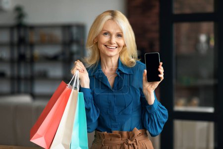 Foto de Mujer madura sonriente demostrando teléfono inteligente en blanco y sosteniendo bolsas de compras brillantes, interior del hogar, maqueta. Feliz hembra recomendando aplicación para compras en línea - Imagen libre de derechos