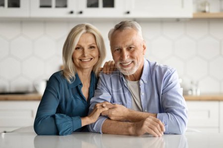 Téléchargez les photos : Portrait de couple de personnes âgées aimant heureux dans des tenues décontractées posant ensemble à l'intérieur de la cuisine confortable, conjoints matures assis à la table, embrasser et sourire à la caméra, profiter du temps à la maison, copier l'espace - en image libre de droit