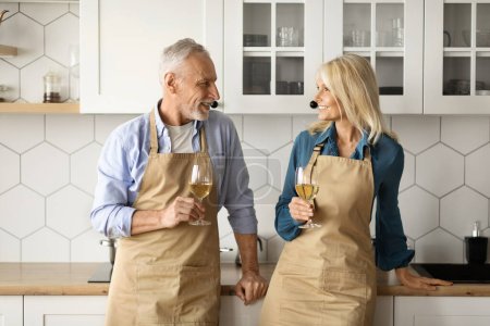 Photo for Portrait Of Romantic Elderly Couple In Aprons Drinking Wine And Relaxing Together In Kitchen While Cooking, Happy Senior Spouses Holding Glasses And Smiling, Enjoying Date At Home, Copy Space - Royalty Free Image