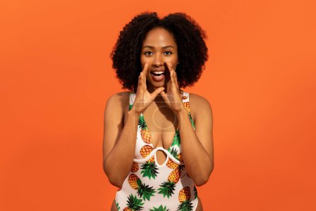 Téléchargez les photos : Offre de voyage. Joyeux attrayant jeune femme noire bouclée en maillot de bain coloré tenant les mains à côté de la bouche et criant bruyamment, faire une annonce sur fond de studio orange, espace de copie - en image libre de droit