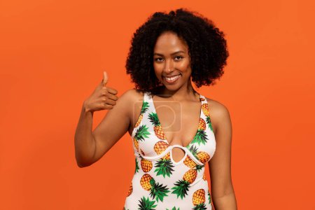 Photo for Positive cheerful pretty millennial black woman with bushy hair in colorful swimwear showing thumb up and smiling isolated on orange studio background, enjoying her summer vacation, holiday - Royalty Free Image