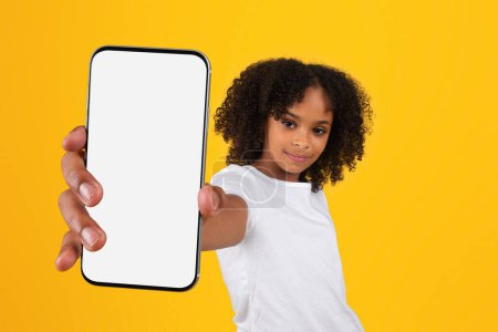 Photo for Cheerful teen curly black girl in white t-shirt show smartphone with blank screen, recommends app, ad, offer, isolated on yellow studio background. Blog and website for education, knowledge study - Royalty Free Image
