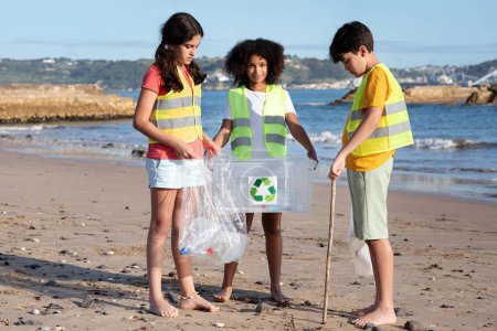 Téléchargez les photos : Des élèves adolescents multiethniques positifs bénévoles en uniforme ramassent les ordures et les bouteilles dans des sacs et des boîtes sur la plage. Sauver l'environnement, nettoyer, protéger la planète, recyclage du plastique - en image libre de droit