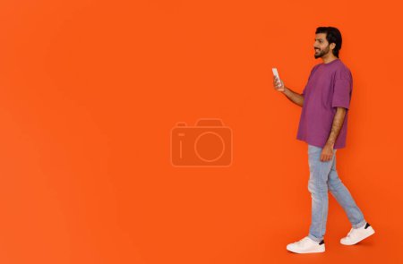 Photo for Cool cheerful handsome young eastern guy in stylish outfit walking with smartphone and smiling on orange studio background, using nice mobile app, panorama with copy space for ad - Royalty Free Image