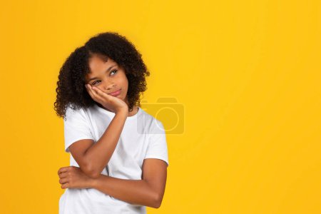 Foto de Triste pensativo cansado adolescente rizado chica en blanco camiseta mira vacío espacio, pensar, aislado amarillo fondo del estudio. Estrés, problemas de estudio, conocimientos en la escuela, emociones de los alumnos - Imagen libre de derechos