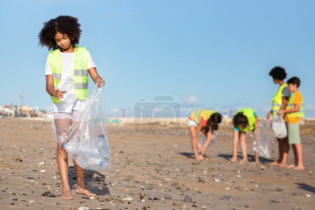 Téléchargez les photos : Divers adolescents occupés en uniforme et fille bouclée noire collectent les ordures et les bouteilles en plastique dans un sac sur la plage. Protection de l'environnement, nettoyage, sauver la planète, recycler, protéger la mer - en image libre de droit