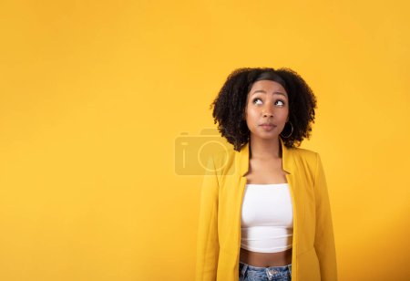 Téléchargez les photos : C'est intéressant. Jeune femme noire incertaine aux cheveux touffus regardant de côté l'espace de copie, posant isolée sur le bakcground studio jaune, bannière - en image libre de droit