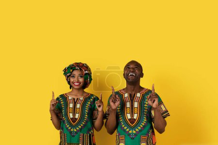 Photo for Positive cheerful bright beautiful african american lovers man and woman in national costumess pointing up at copy space above their heads, showing nice advertisement, yellow background - Royalty Free Image