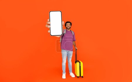 Foto de Alegre guapo joven indio turista mostrando gran teléfono inteligente con pantalla blanca vacía, aislado en fondo de estudio naranja. Check-in en línea, reserva de vuelos, aplicación móvil itinerante, maqueta - Imagen libre de derechos