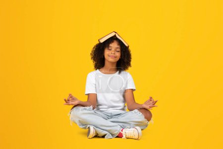 Téléchargez les photos : Heureuse adolescente afro-américaine frisée en t-shirt blanc avec livre sur la tête et les yeux fermés, se repose, méditant en position lotus, isolée sur fond de studio jaune. Pause, lecture, étude et connaissance - en image libre de droit