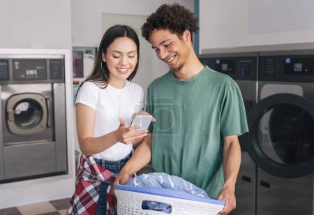 Photo for Cheerful Young Spouses Booking Laundry Online Via Application On Smartphone Standing Near Washing Machines, Posing With Basket Full Of Clothes Indoors. Technology And Laundrette Chores Concept - Royalty Free Image
