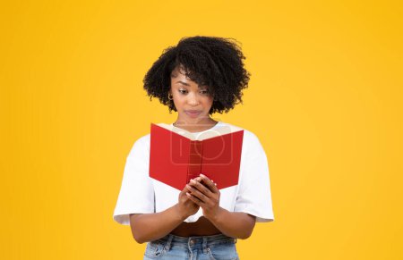 Téléchargez les photos : Joyeux sérieux millénium intelligent afro-américaine étudiante frisée en blanc livre de lecture de t-shirt, isolé sur fond de studio jaune. Études, devoirs, apprentissage et connaissances - en image libre de droit