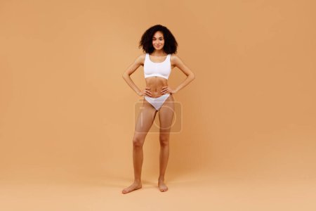 Téléchargez les photos : Joyeux jeune dame mince posant en sous-vêtements blancs et montrant ses belles courbes et sa forme corporelle parfaite sur fond de studio beige, espace de copie, plan complet - en image libre de droit