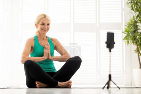 Téléchargez les photos : Cours de yoga en ligne. Femme âgée souriante Méditant devant Smartphone à la maison, Femme mûre sportive assise dans Lotus Pose, capturant du contenu pour les médias sociaux ou assistant à une formation virtuelle - en image libre de droit