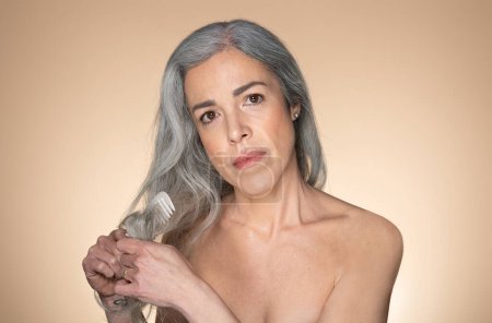 Téléchargez les photos : Femme aux cheveux gris sénior grave peigner ses cheveux avec peigne, isolé sur fond studio beige, espace de copie. Soins de beauté à la maison procédure quotidienne, traitements et problèmes de cheveux - en image libre de droit