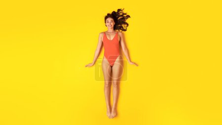 Photo for Ready For Summer Vacation. Happy Lady In Orange Swimwear Posing Smiling To Camera Lying On Yellow Studio Background, Panorama, View From Above. Slim Woman Sunbathing And Relaxing Wearing Swimsuit - Royalty Free Image