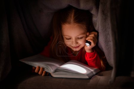 Téléchargez les photos : Petite fille souriante lisant le livre tout en se cachant sous la couverture avec la lampe de poche, enfant féminin mignon joyeux tenant la torche de poche dans l'obscurité et appréciant ses Tailes Faily préférés, gros plan tiré - en image libre de droit