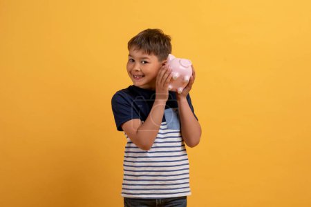 Photo for Happy boy holding piggy bank for savings, money or coins next to ear against yellow studio background. Little child or kid checking piggybank and smiling for financial freedom, cash or investment - Royalty Free Image