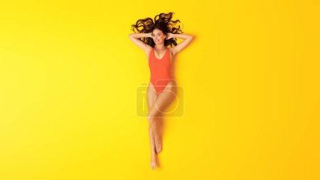 Photo for Summer Vacation. Young Woman In Orange One Piece Swimsuit Lying Sunbathing And Relaxing Over Yellow Studio Background, Smiling To Camera. Top View Shot Of Lady Wearing Swimwear. Panorama - Royalty Free Image