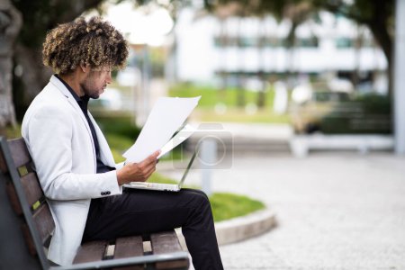 Téléchargez les photos : Concept de carrière en affaires. Entrepreneur afro-américain d'âge moyen travaillant sur un ordinateur portable et lisant des documents, assis sur un banc à l'extérieur, vue latérale, espace de copie - en image libre de droit