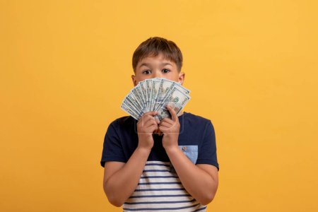 Photo for Kids and financial letiracy cocnept. Amazed cute rich boy school aged holding bunch of cash dollar banknotes, covering face with money, isolated on yellow studio background - Royalty Free Image
