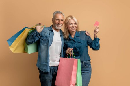 Photo for Positive happy beautiful elderly couple man and woman wearing denim outfits posing with colorful shopping bags, showing bank card over beige studio background. Contactless payment, unlimited shopping - Royalty Free Image