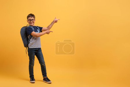 Photo for Happy school boy wearing glasses, backpack and casual outfit posing on yellow studio background, smart schooler going to school, enjoying studying, pointing at copy space. School, education concept - Royalty Free Image