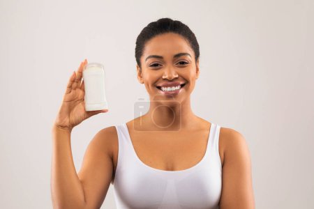 Foto de Joven afro-americana alegre y positiva con la parte superior blanca sosteniendo desodorante en la mano y sonriendo en el fondo gris del estudio, espacio para copiar. Cuidado corporal, axilas, concepto de higiene de axilas - Imagen libre de derechos