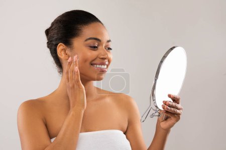 Photo for Skin care routine. Happy attractive african american millennial woman looking at small mirror, massaging face with fingers and smiling, applying cream, serum or oil, isolated on grey, copy space - Royalty Free Image