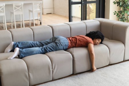 Foto de Mujer negra joven y cansada durmiendo en el sofá durante el día, sintiéndose agotada, milenaria dama afroamericana durmiendo la siesta en casa, relajándose en un acogedor sofá con la cara hacia abajo, disfrutando de un fin de semana perezoso, espacio para copiar - Imagen libre de derechos