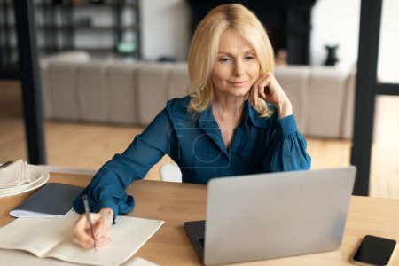 Photo for Attractive mature businesswoman making notes at table with laptop, watching online lesson or webinar at home office interior. Work and business remotely - Royalty Free Image
