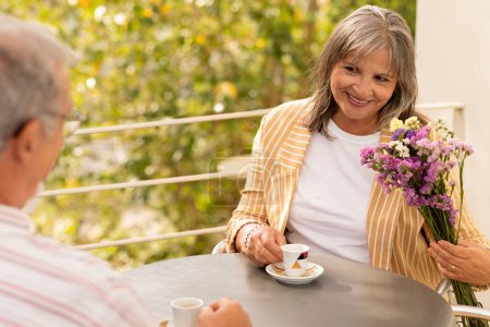 Téléchargez les photos : Heureuse femme caucasienne âgée avec bouquet regardez l'homme, buvez du café, asseyez-vous à table dans un café en plein air. Couple profiter de romance, moment tendre, premier rendez-vous et mode de vie actif, amour et relation - en image libre de droit