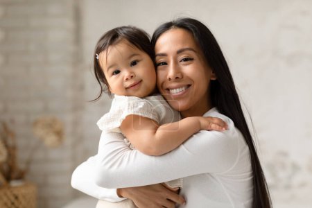 Téléchargez les photos : Faire place à l'amour. Asiatique maman tendrement étreignant et tenant sa petite fille enfant dans la chambre à coucher, souriant à la caméra. Fiers mère posant avec bébé fille mignonne à la maison, coller et profiter du temps ensemble - en image libre de droit