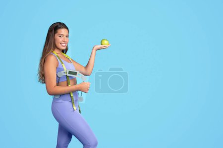 Photo for Smiling millennial caucasian slim woman in sportswear with scales and measuring tape, show green apple, isolated on blue studio background. Sports, slimming and body care, diet, ad and offer - Royalty Free Image