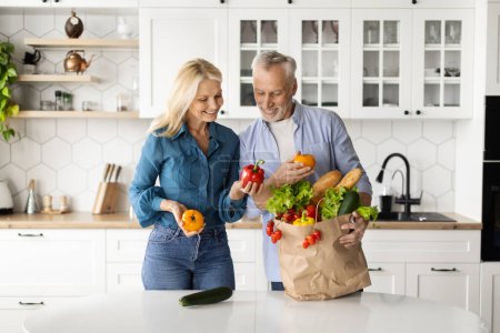 Téléchargez les photos : Livraison de nourriture biologique. heureux couple aîné déballer sac avec épicerie dans la cuisine, joyeux aînés conjoints tenant légumes frais et souriant, mari et femme mûrs jouissant d'une alimentation saine - en image libre de droit