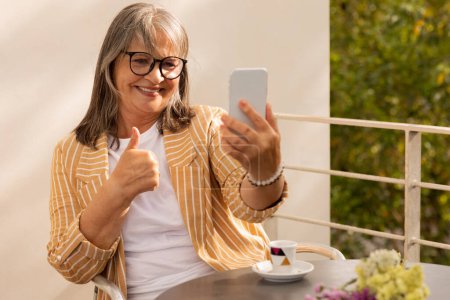 Foto de Feliz vieja caucásica de pelo gris mujer en gafas se sienta a la mesa en la cafetería, hacer selfie, tiene videollamada en el teléfono inteligente, mostrar el pulgar hacia arriba gesto, al aire libre. Lady disfruta de la aplicación, descanso, comunicación de forma remota - Imagen libre de derechos